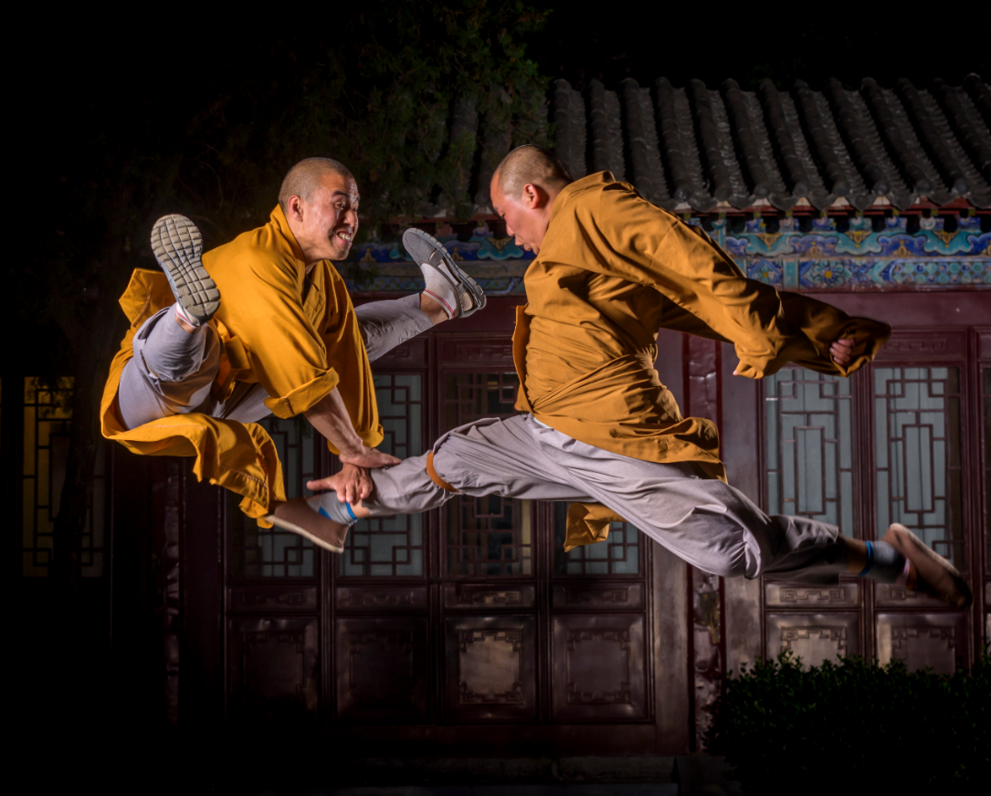 Two Shaolin monks performing high-flying kung fu kicks in traditional robes outside a temple.