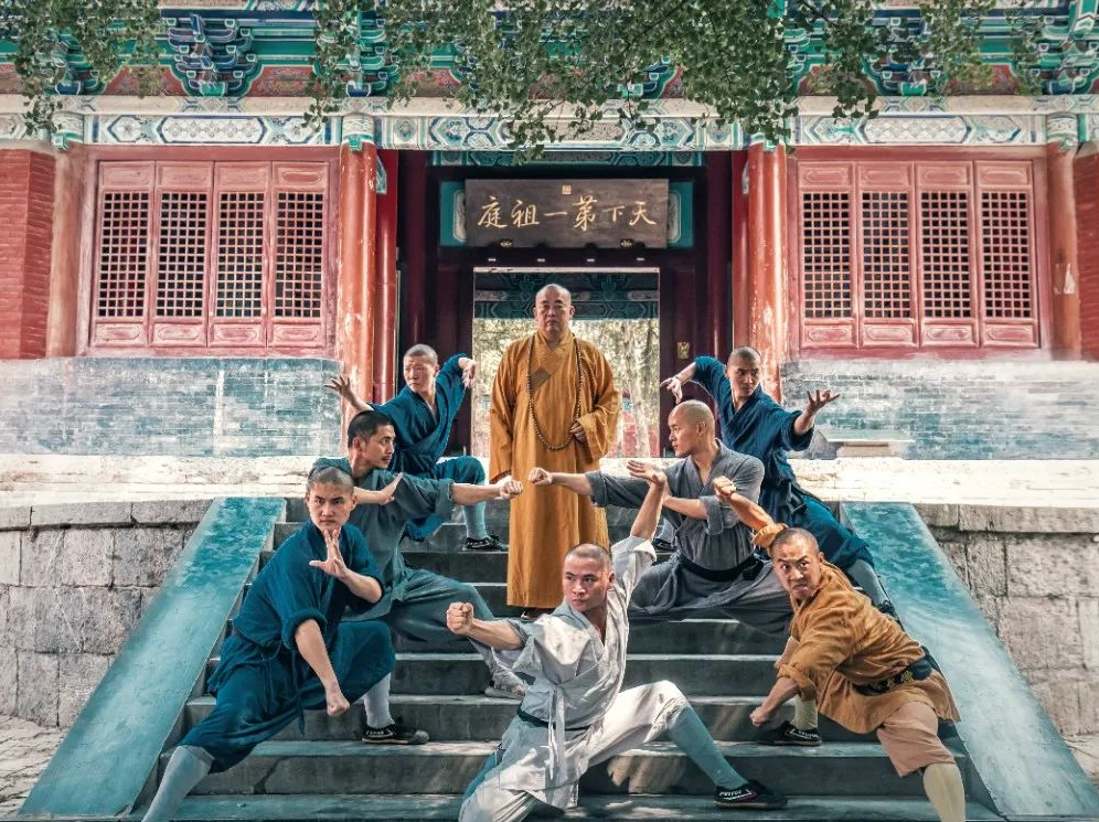 Shaolin monks in traditional robes pose in martial arts stances at a Buddhist temple entrance.