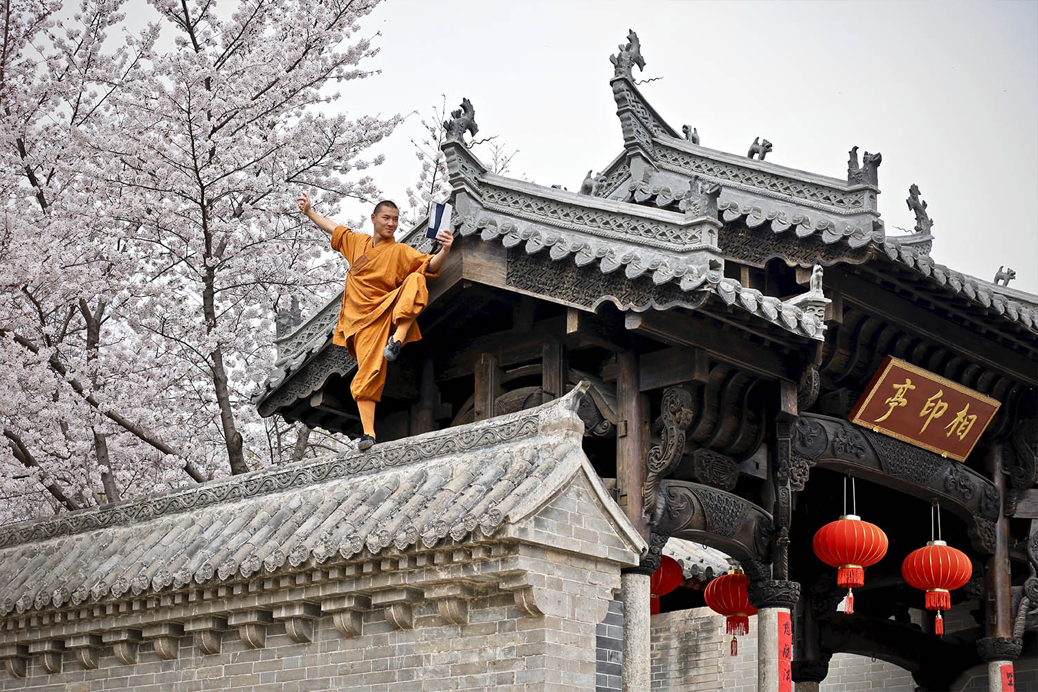 Shaolin monk in orange robe balancing on traditional temple roof, cherry blossoms and red lanterns