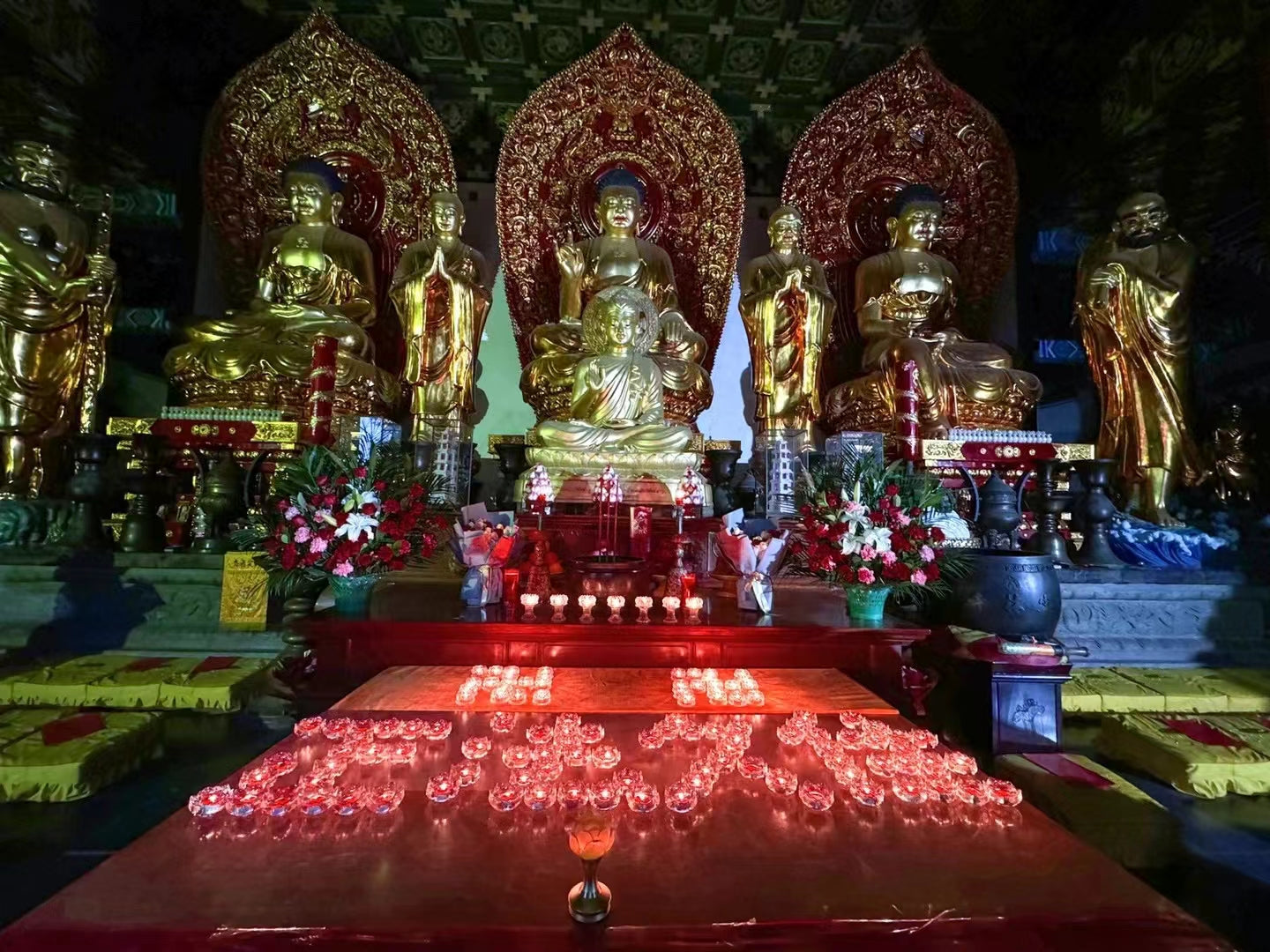 Golden Buddhist statues on ornate altar with red candles and prayer cushions in temple setting