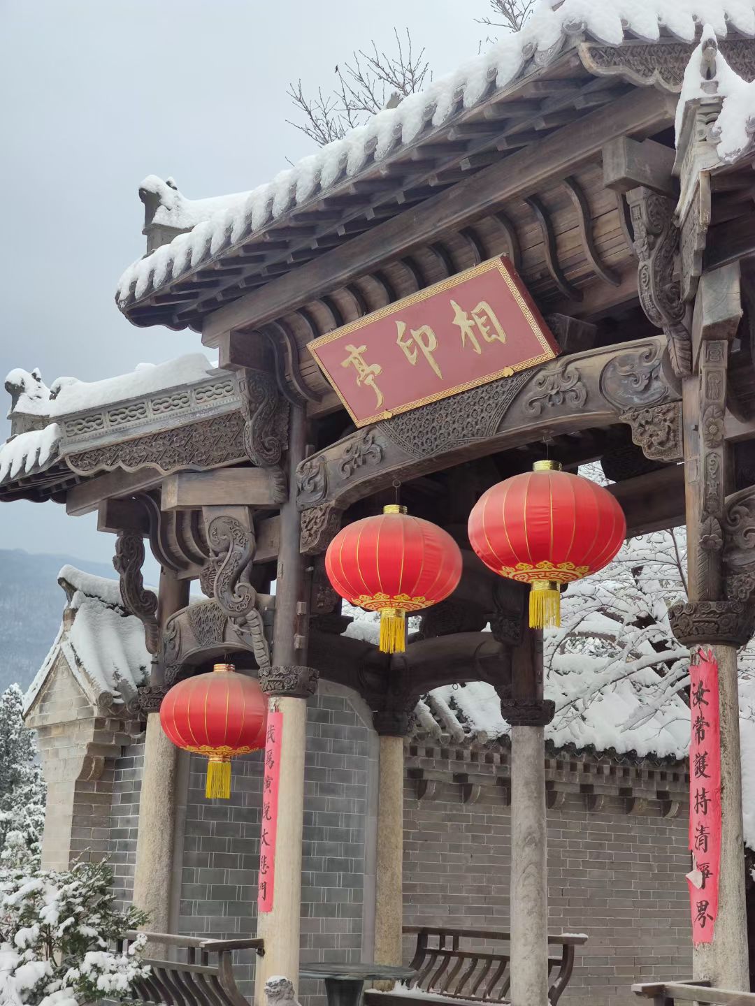 Traditional Chinese temple gate with red lanterns and snow-covered roof in winter.