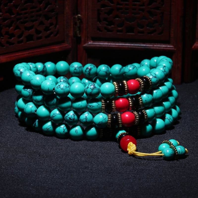 Turquoise prayer beads with red guru beads, black spacers, and gold accents, coiled on dark fabric near carved wood