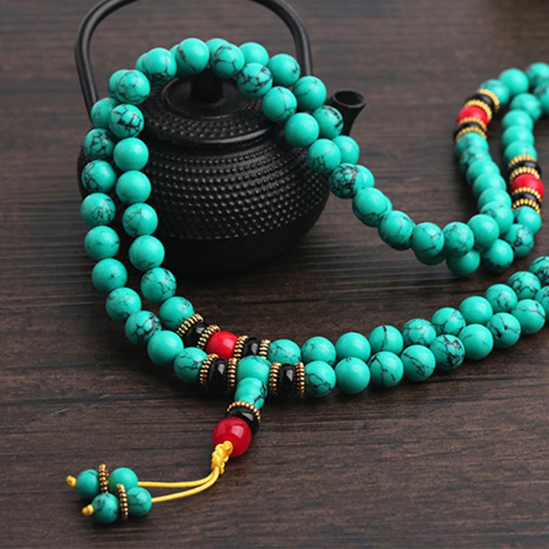 Turquoise Buddhist prayer beads with red and black accents, displayed near a black teapot on wood.