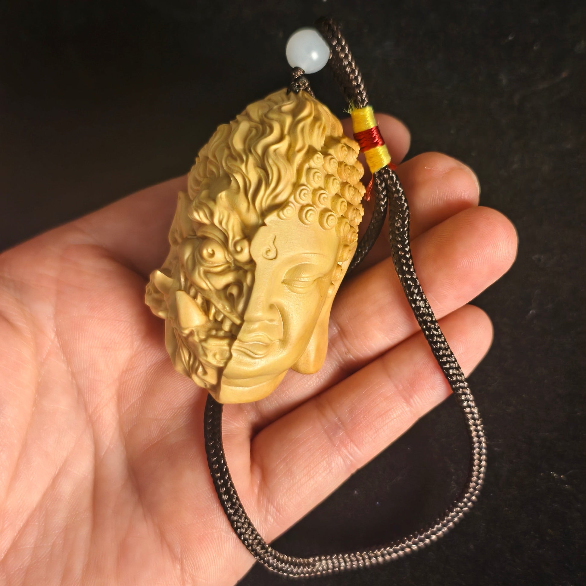 Hand holding carved Buddha head pendant with dual serene and fierce faces, on braided cord with jade bead