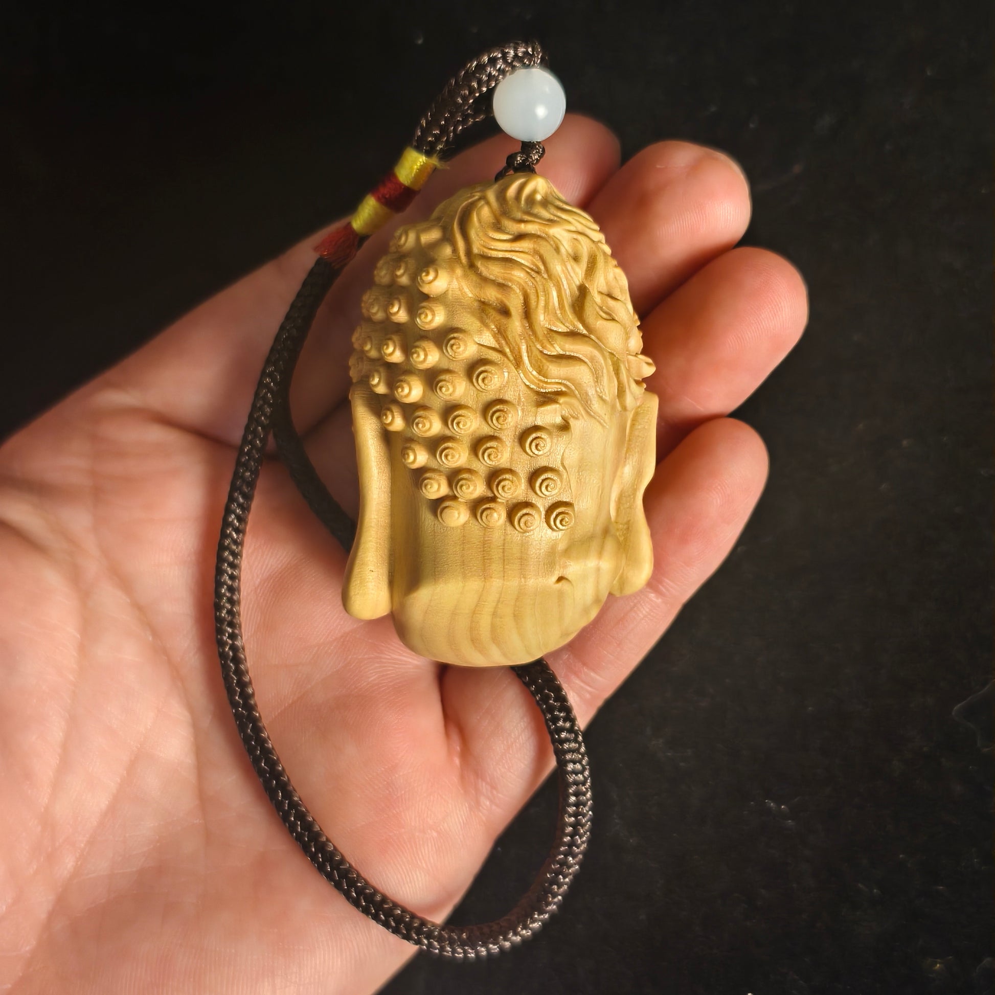 Hand-carved wooden Buddha head pendant with spiral curls, on braided cord with jade bead, for mindfulness and peace.