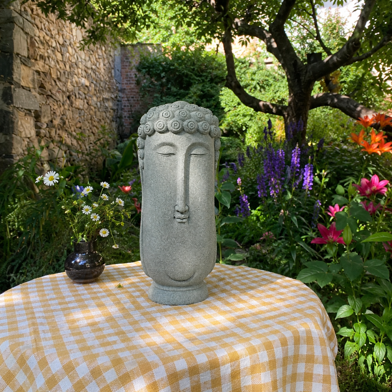 Stone Buddha head statue on checkered tablecloth in garden, surrounded by flowers and greenery for mindful decor