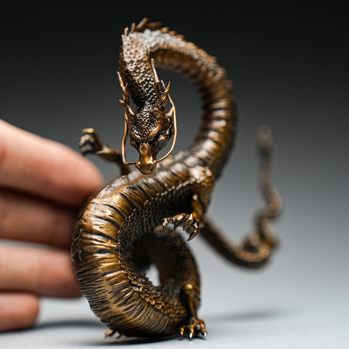 Bronze-colored Chinese dragon statue with intricate scales, held by a hand, on gray background