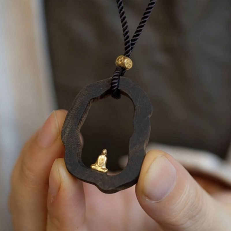 Hand holding dark wood pendant with gold Buddha statue, black cord necklace for mindfulness and peace