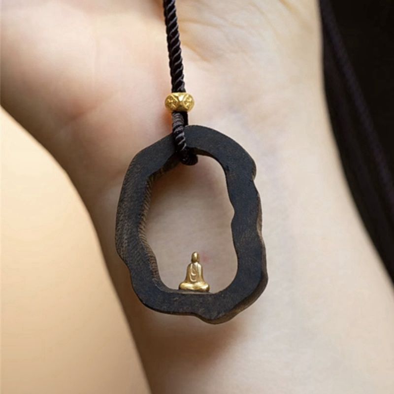 Hand holding dark wood pendant with gold Buddha statue, black cord necklace for mindfulness and peace